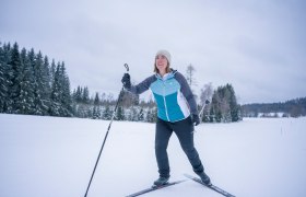 Eine Frau beim Langlaufen auf einer verschneiten Landschaft mit B&auml;umen im Hintergrund.