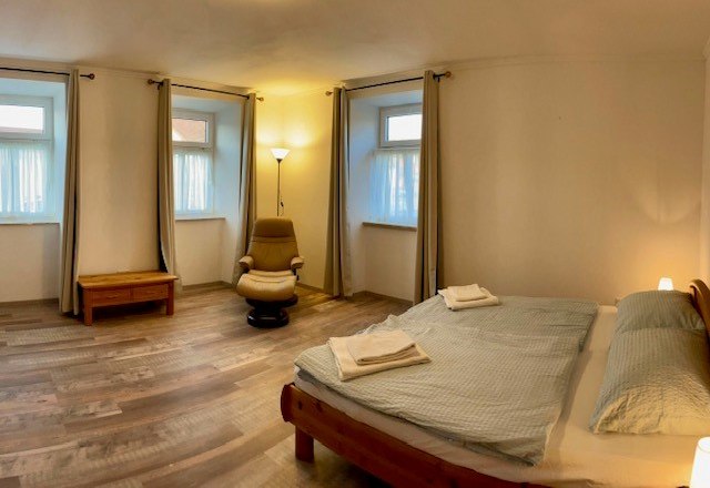 Ein schlichtes Schlafzimmer mit Doppelbett, Sessel, Nachttisch und drei Fenstern mit Vorhängen.