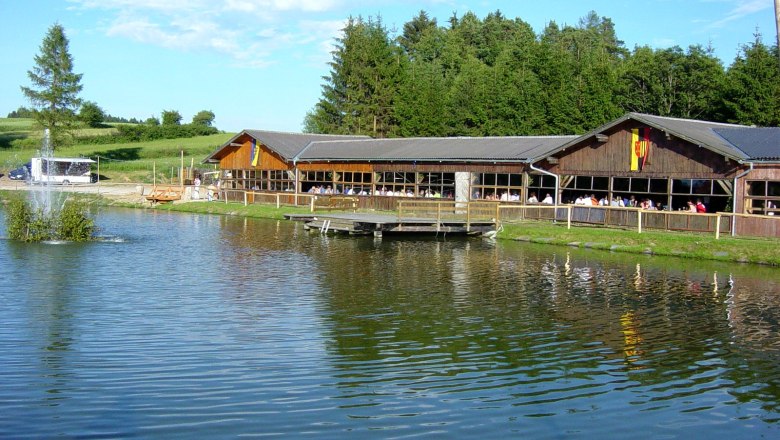 Ein Holzgeb&auml;ude an einem Teich mit Springbrunnen, umgeben von B&auml;umen und Wiesen.