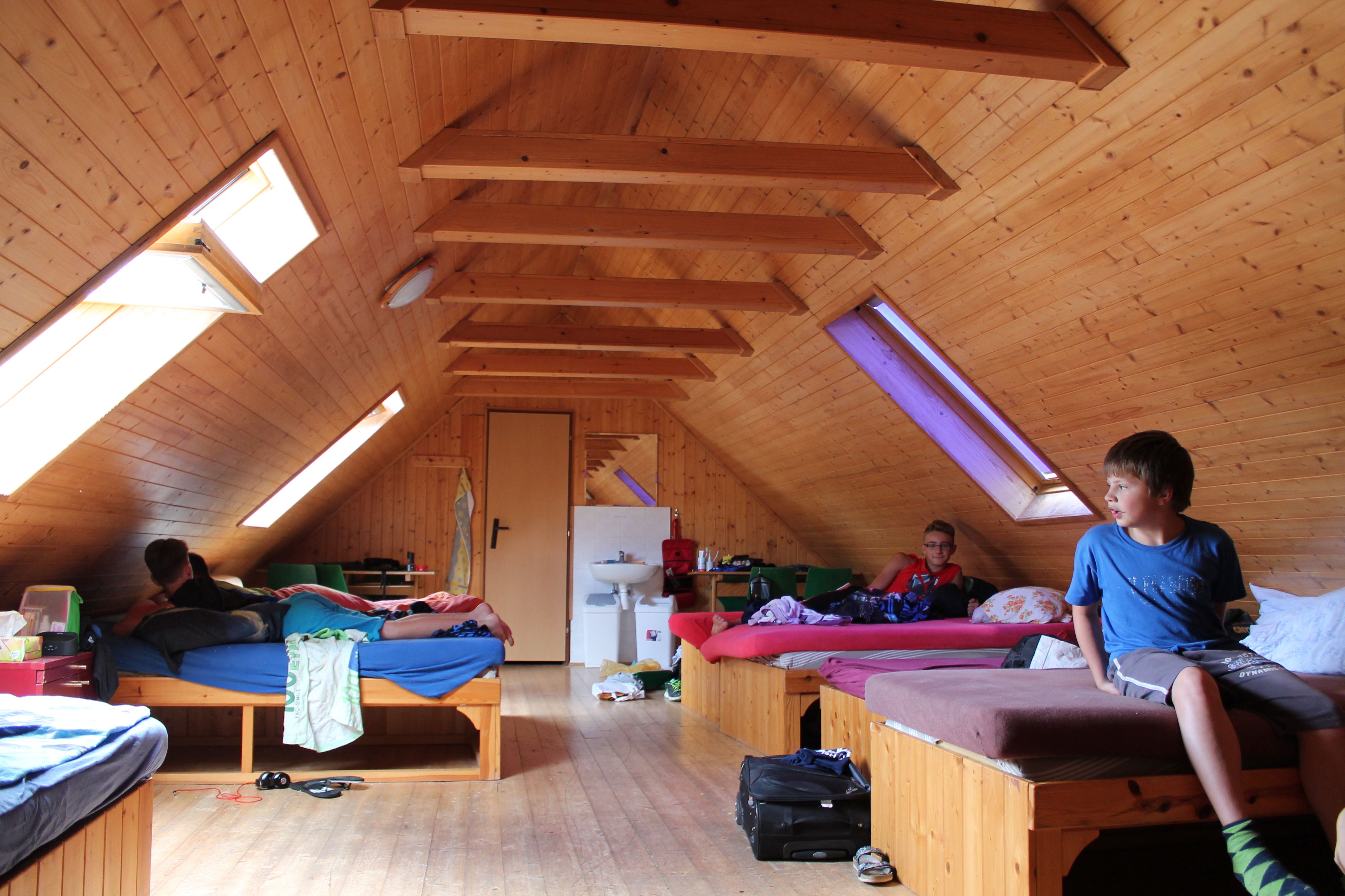 Dachgeschosszimmer mit Holzwänden, mehreren Betten und Dachfenstern. Drei Personen sind im Raum, eine sitzt auf einem Bett.