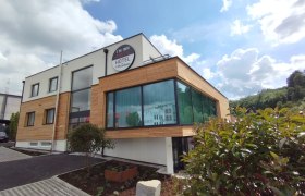 Modernes Hotelgebäude mit Holzverkleidung und großen Fenstern, umgeben von Pflanzen, unter blauem Himmel mit Wolken.