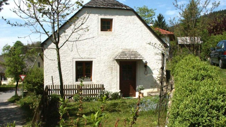 Kleines weißes Ferienhaus mit Garten und Bäumen, umgeben von grüner Landschaft.
