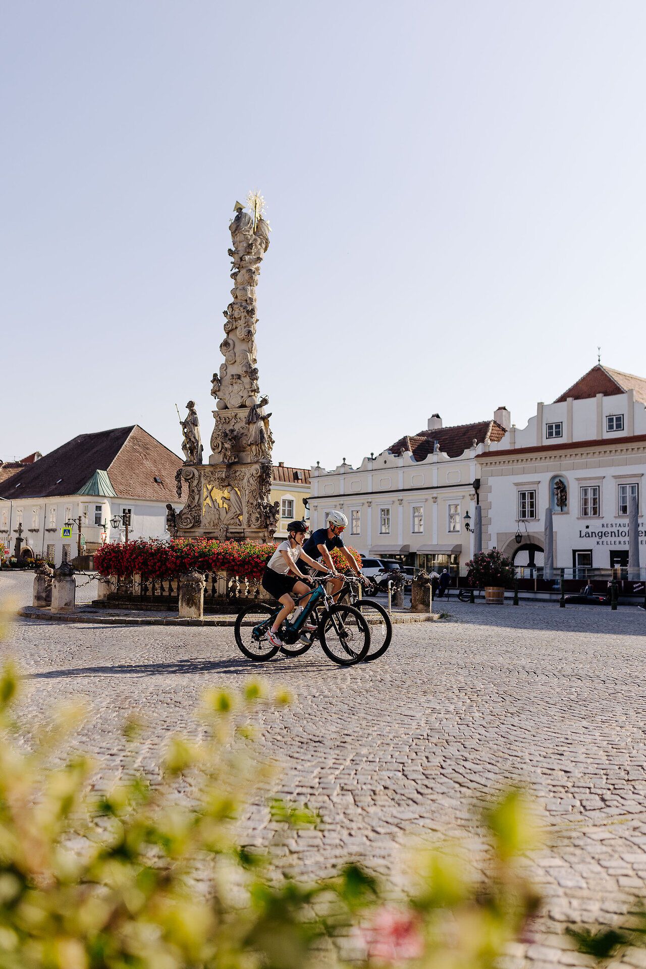 Zwei Radfahrer auf ihren Rädern fahren über den Hauptplatz von Langenlois.