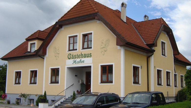 Gästehaus Hahn, © Gästehaus Hahn Gelbes Gästehaus mit rotem Dach und geparkten Autos davor.