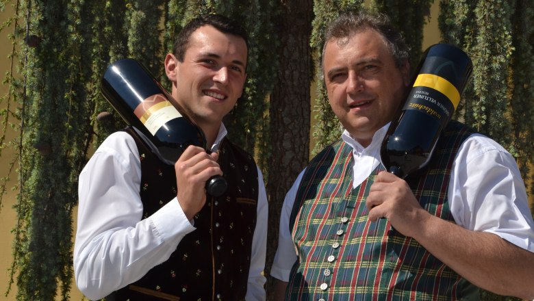 Zwei Männer in traditioneller Kleidung halten große Weinflaschen vor einem Baum.