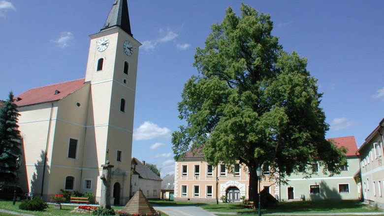Kirche mit Turm und Uhr in einem Dorfplatz mit Bäumen und Gebäuden.
