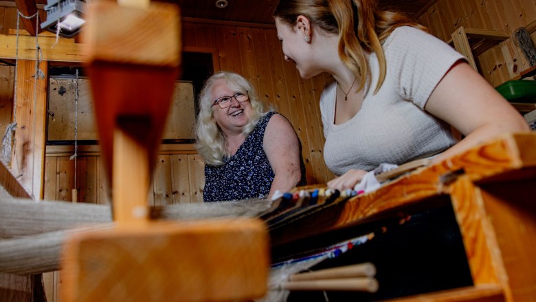 Zwei Frauen beim Weben eines Fleckerlteppichs in einem holzgetäfelten Raum.