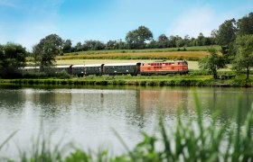 Ein Zug fährt entlang eines Flusses durch eine grüne Landschaft unter blauem Himmel.