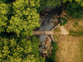Zwei Personen kommen aus einem Waldstück und gehen über eine Brücke zu einer Wiese