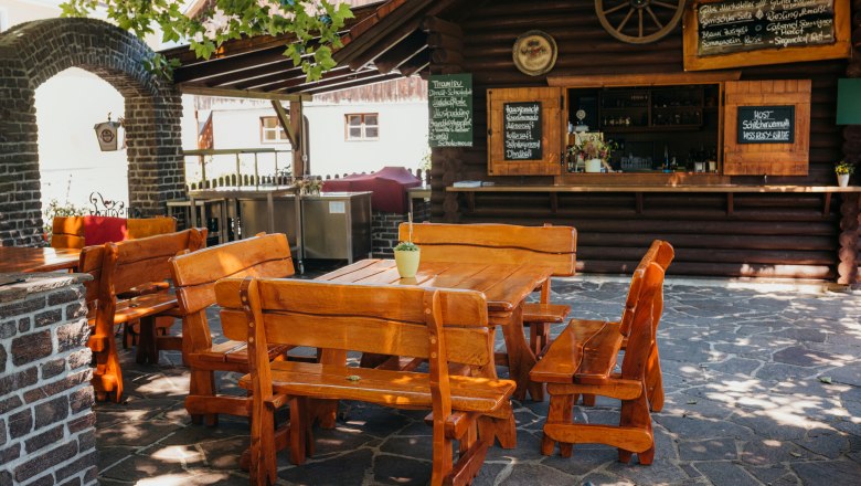 Rustikaler Gastgarten mit Holztischen und -b&auml;nken, umgeben von Steinmauern und einem kleinen Barbereich.