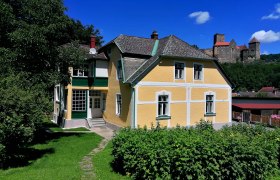 Gelbes Haus mit grünem Garten, im Hintergrund eine Burg auf einem Hügel.