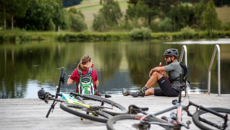 Zwei Radfahrer sitzen auf einem Steg am Frauenwieserteich, umgeben von Fahrrädern und Natur.