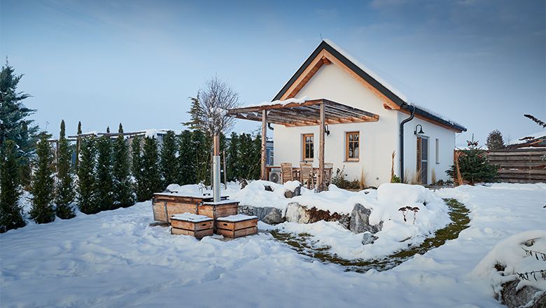 Ein Chalet im Schnee mit Terrasse und Gartenmöbeln, umgeben von Bäumen.