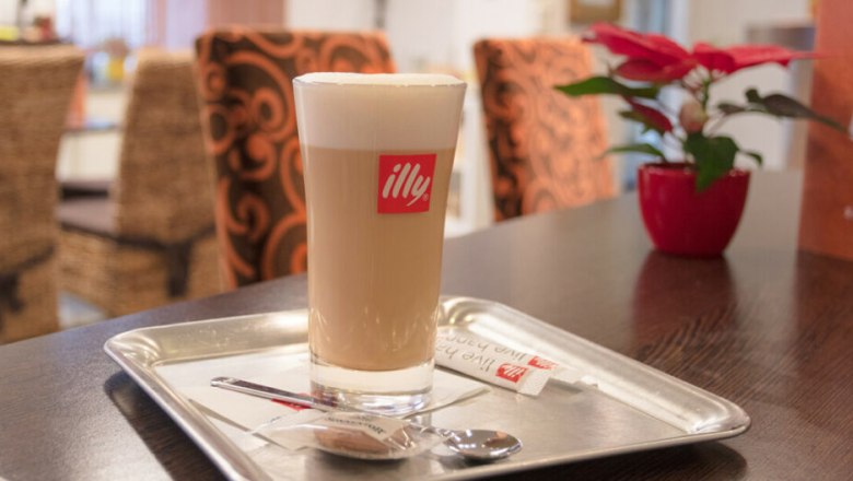 Ein Glas Caf&eacute; Latte auf einem Tablett mit einem L&ouml;ffel und Zucker, im Hintergrund ein roter Blumentopf auf einem Tisch.