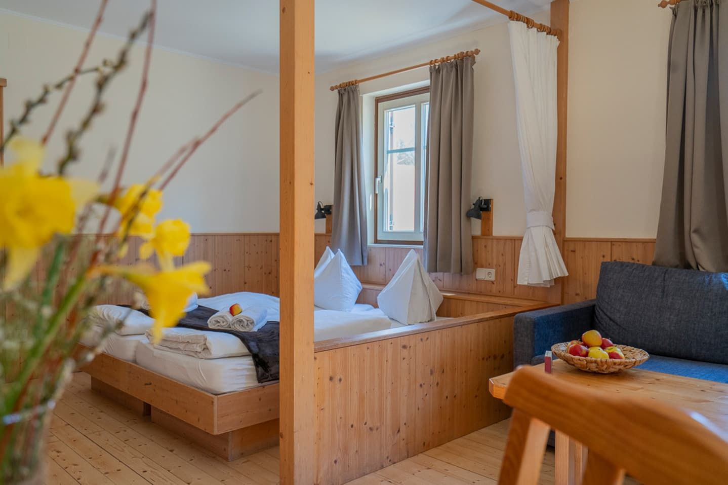 Gemütliches Zimmer mit Holzmöbeln, einem Bett, einem Sofa und einem Tisch mit Obstschale. Gelbe Blumen im Vordergrund.