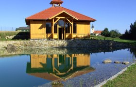 Holzgebäude mit rotem Dach spiegelt sich in einem Teich, umgeben von grüner Landschaft und blauem Himmel.