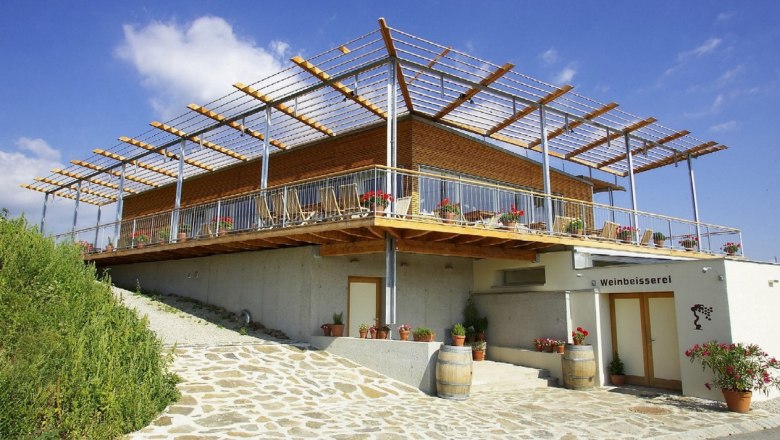 Modernes Gebäude mit Terrasse und Pergola, umgeben von Pflanzen und Weinfässern, unter blauem Himmel.