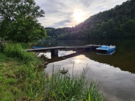 Ein ruhiger Stausee mit einem Steg und einem Tretboot im Wasser, umgeben von gr&uuml;nen B&auml;umen und bew&ouml;lktem Himmel.