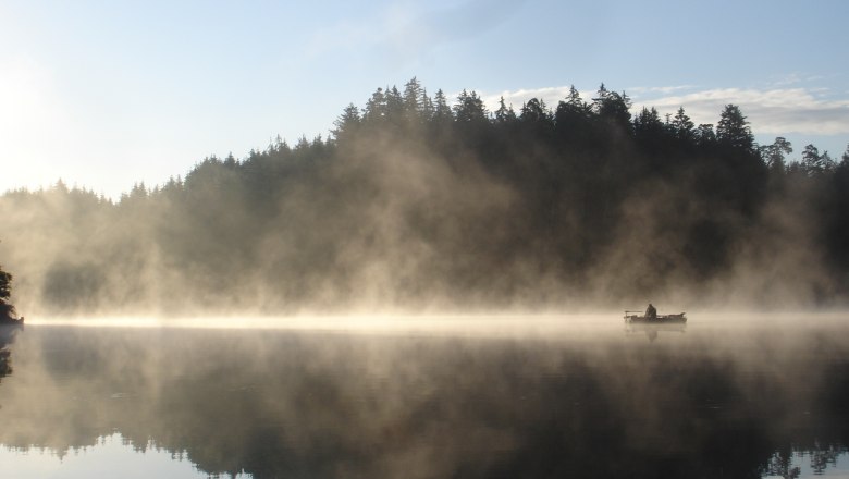 Ein Boot auf einem nebligen See vor einem bewaldeten Ufer bei Sonnenaufgang.