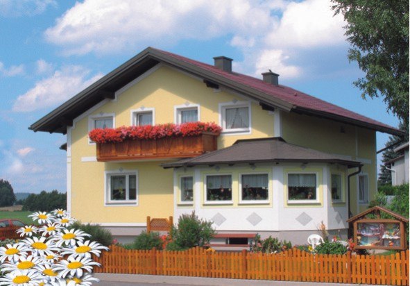 Ein gelbes Haus mit rotem Dach und Blumenkasten auf dem Balkon, umgeben von einem Holzzaun und Gänseblümchen im Vordergrund.