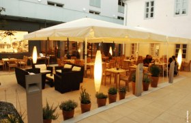 An illuminated guest garden with tables, chairs and plants in pots, surrounded by buildings.