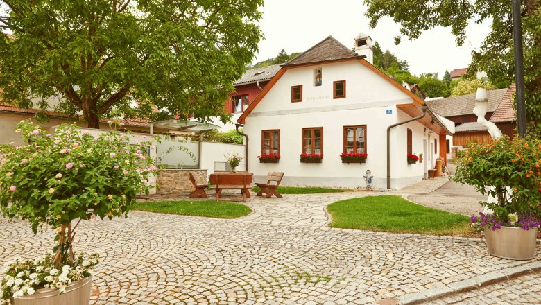 Ein malerischer Platz mit Kopfsteinpflaster, einem kleinen weißen Haus mit roten Fensterläden und Blumen, umgeben von Bäumen und Pflanzen.