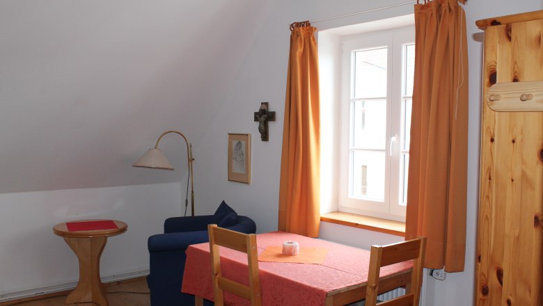 Gemütliches Zimmer mit Tisch, Stühlen, Sofa und Fenster mit orangefarbenen Vorhängen.