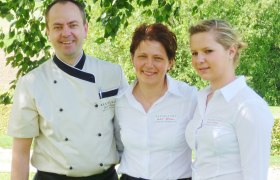 Drei Personen in Restaurantuniformen stehen lächelnd unter einem Baum.