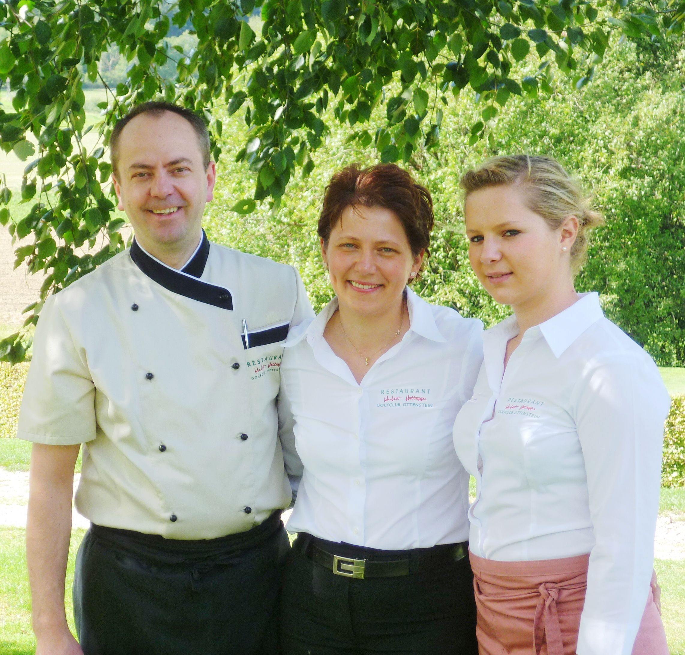 Drei Personen in Restaurantuniformen stehen lächelnd unter einem Baum.