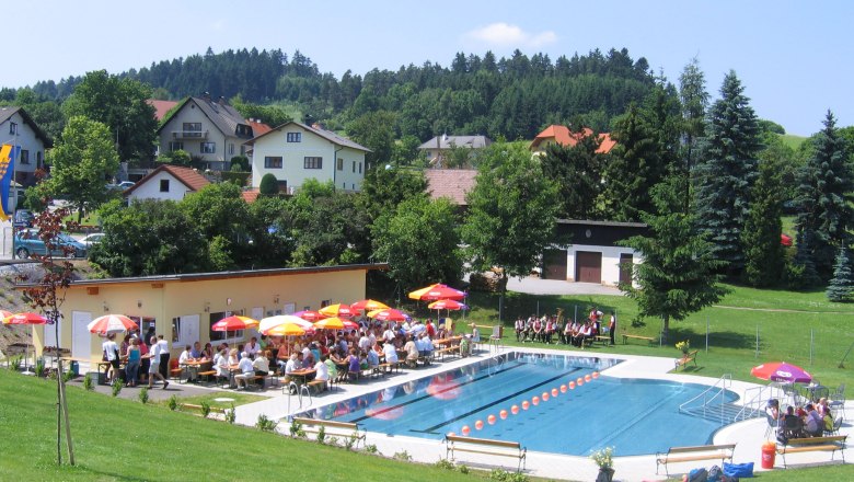 Freibad mit Schwimmbecken, Sonnenschirmen und Menschen auf Liegest&uuml;hlen, umgeben von gr&uuml;nen H&uuml;geln und H&auml;usern.