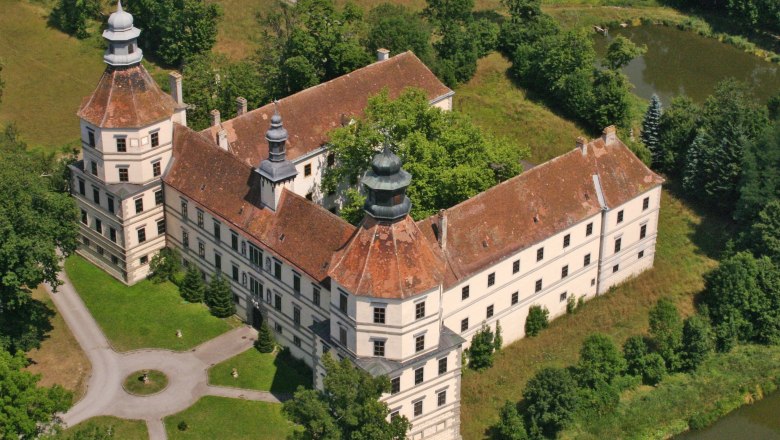 Luftaufnahme eines historischen Schlosses mit zwei Türmen und einem Innenhof, umgeben von Bäumen und einem Teich.
