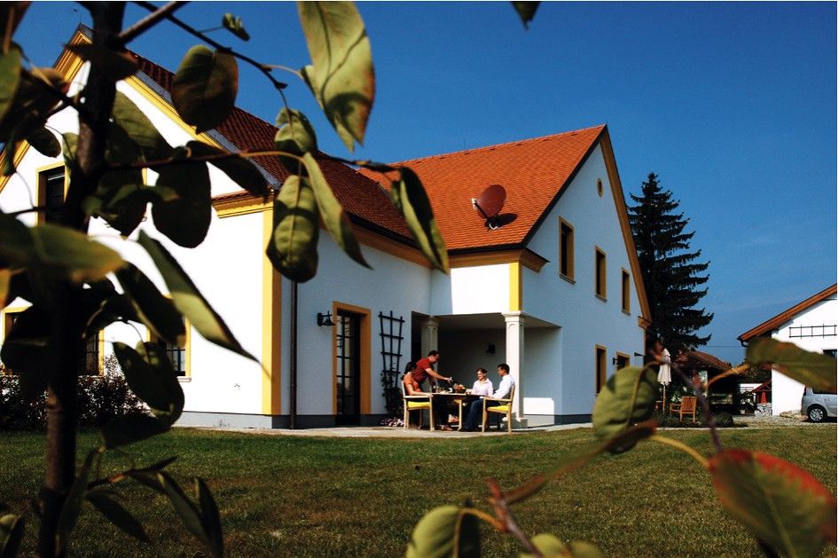 Ein weißes Haus mit rotem Dach, umgeben von grünem Rasen und Bäumen. Menschen sitzen an einem Tisch im Freien.