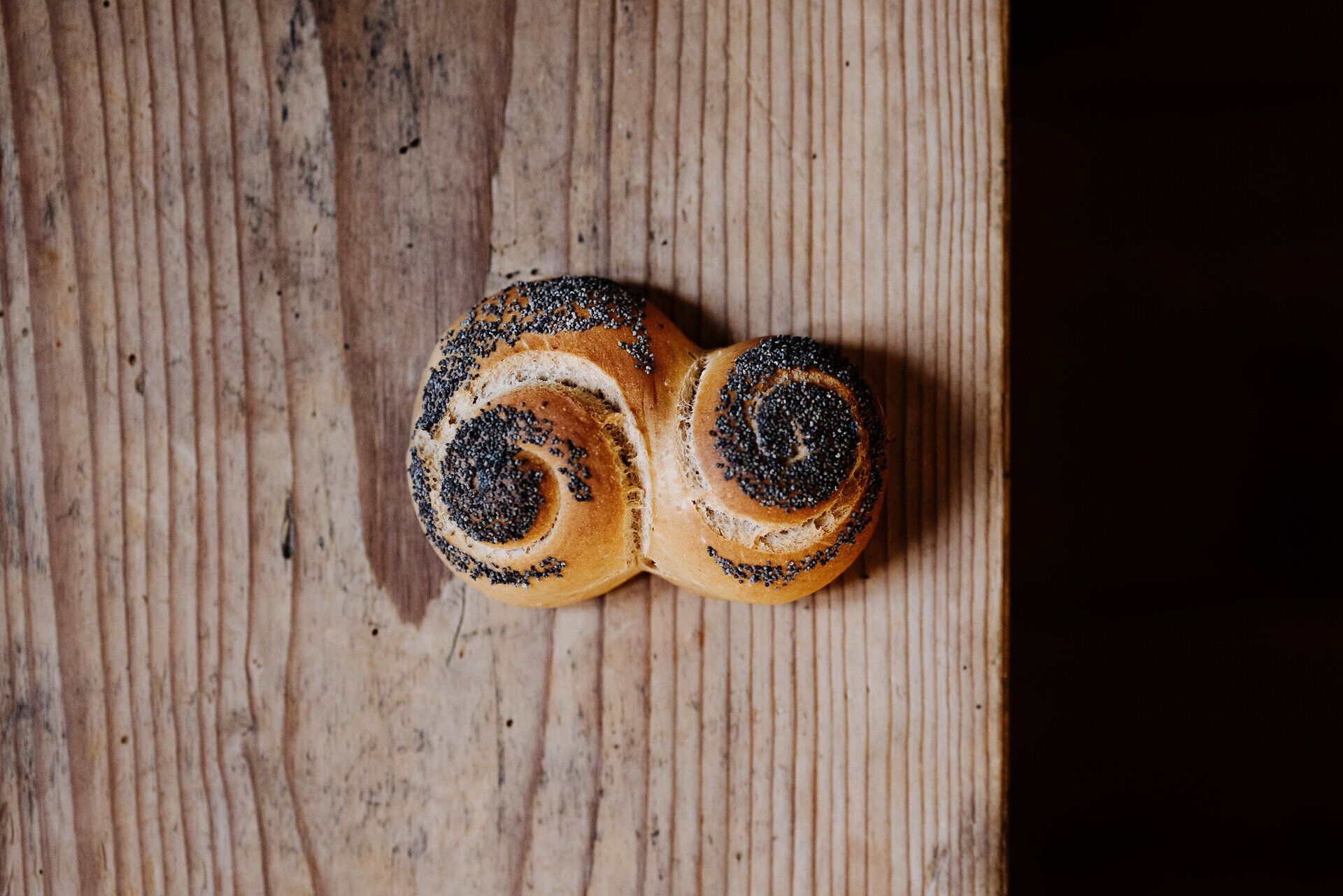 Ein frisch gebackenes Mohnweckerl liegt verführerisch auf einem rustikalen Holztisch. Der Duft von warmem Brot und die knusprige Textur laden dazu ein, sofort hinein zu beißen und die Aromen zu genießen. Hier in der Bio-Backschule wird Tradition mit Liebe zum Detail gelebt.