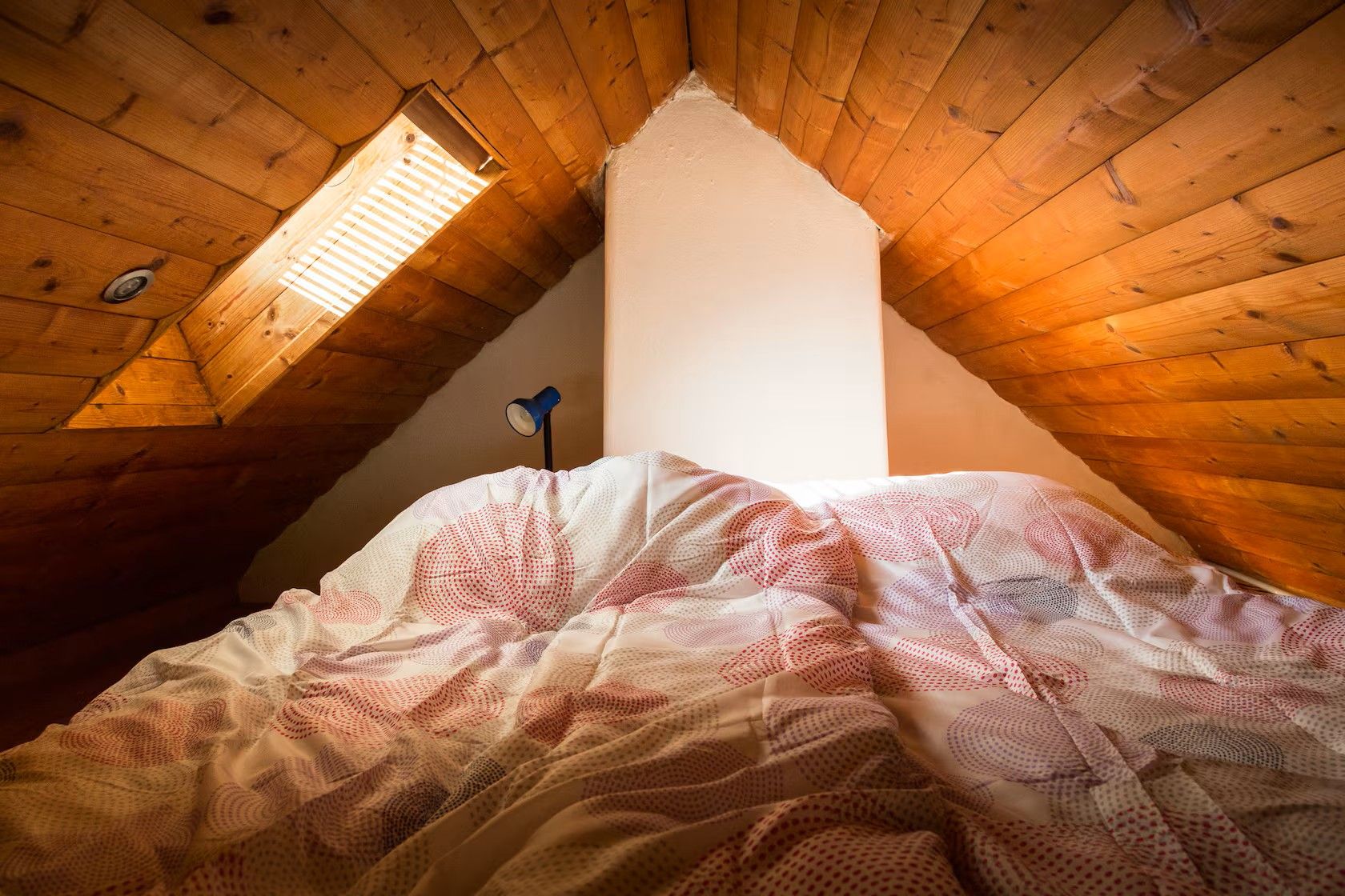 Dachzimmer mit Holzdecke, Dachfenster und ungemachtem Bett mit gemusterter Bettdecke.