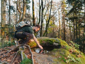 In der malerischen Ysperklamm, umgeben von majest&auml;tischen B&auml;umen und sanften H&uuml;geln, kniet ein Wanderer am Ufer eines klaren Baches. Die goldenen Herbstbl&auml;tter reflektieren das sanfte Licht der Nachmittagssonne und schaffen eine friedliche Atmosph&auml;re, die zum Verweilen einl&auml;dt.