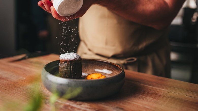 Person bestreut Schokoladenkuchen mit Puderzucker, daneben eine Mandarine auf einem Teller.