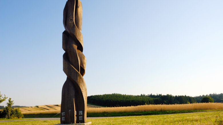Holzskulptur auf einem Feld mit blauem Himmel im Hintergrund.