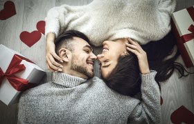 Ein Paar liegt lächelnd auf dem Boden, umgeben von roten Herzen und Geschenken mit roten Schleifen.