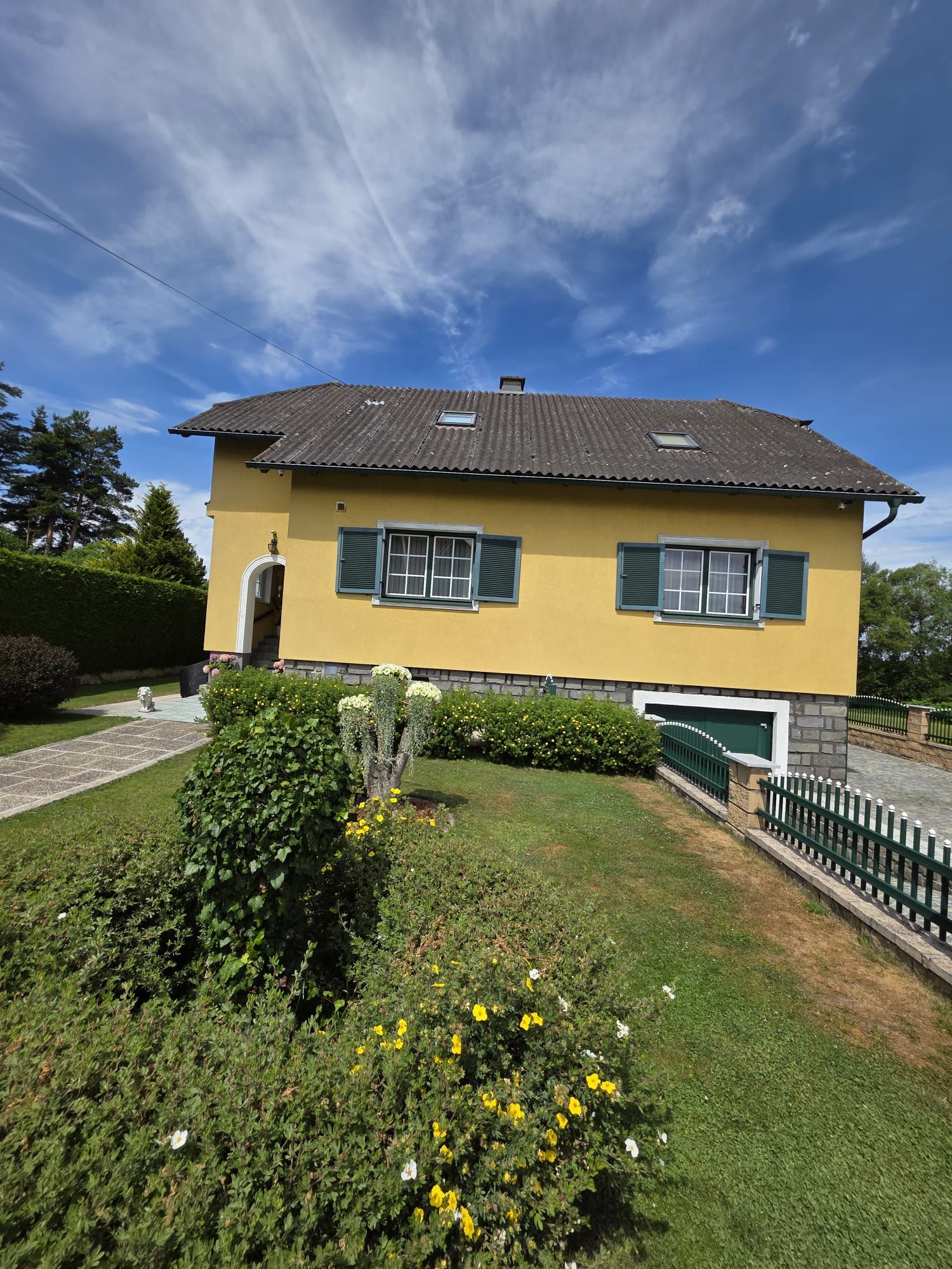 Gelbes Haus mit grünen Fensterläden und gepflegtem Garten unter blauem Himmel.
