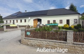 Ein großes Gebäude mit gelber Fassade und schwarzem Dach, umgeben von einem Garten und einem Holzzaun. Der Himmel ist blau mit einigen Wolken.