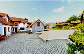 Hof mit mehreren Geb&auml;uden, blauer Himmel, gepflasterter Boden, Blumen und Pflanzen.