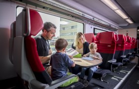 Eine Familie sitzt in einem Zugabteil der ÖBB und beschäftigt sich mit Büchern und Spielen.