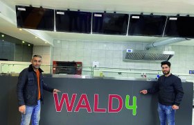 Zwei Männer stehen vor einem Tresen mit der Aufschrift 'WALD4' in einer Pizzeria.