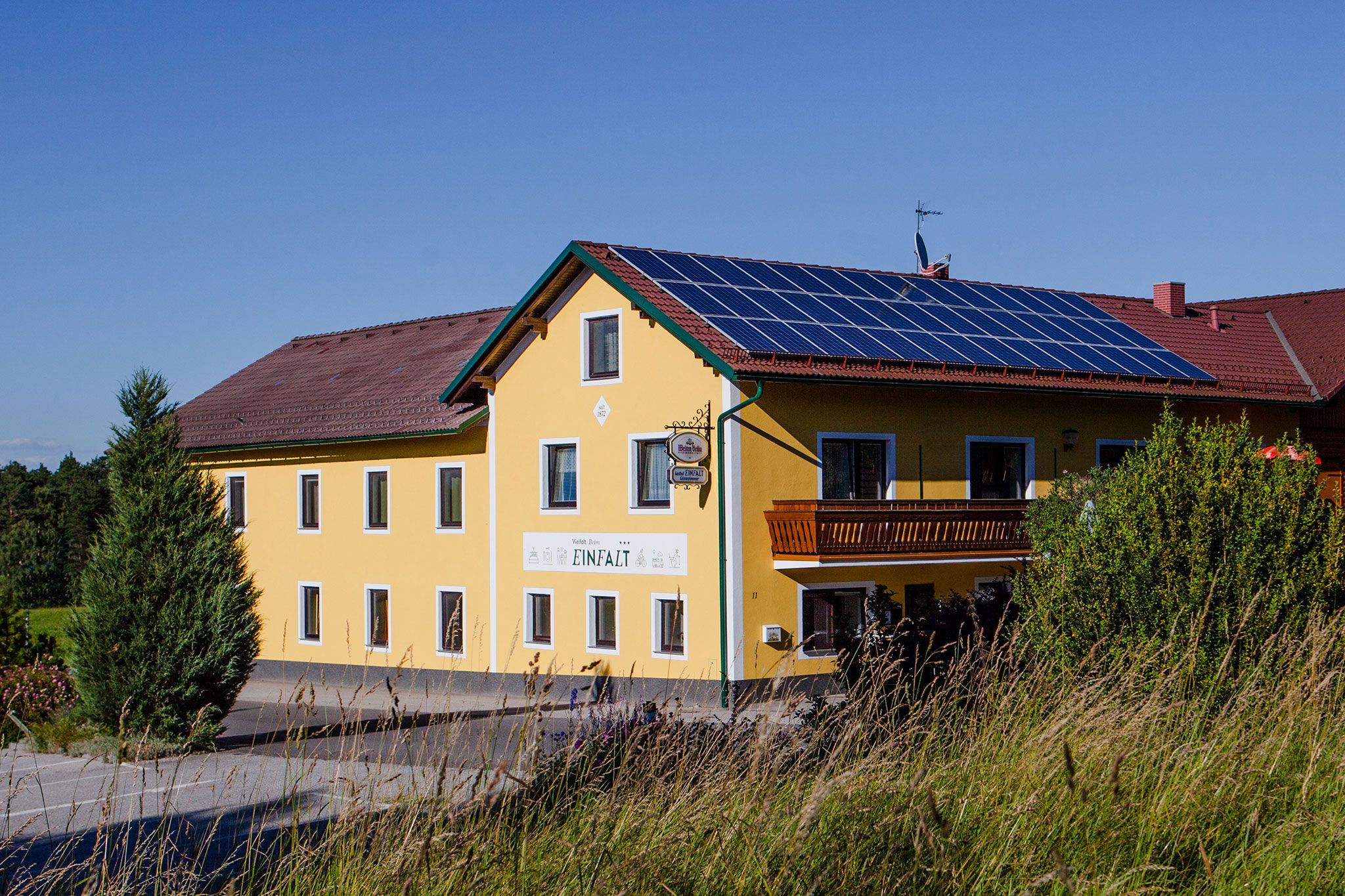 Gelbes Gasthaus mit Solarpanelen auf dem Dach und einem Balkon, umgeben von Bäumen und Gras.
