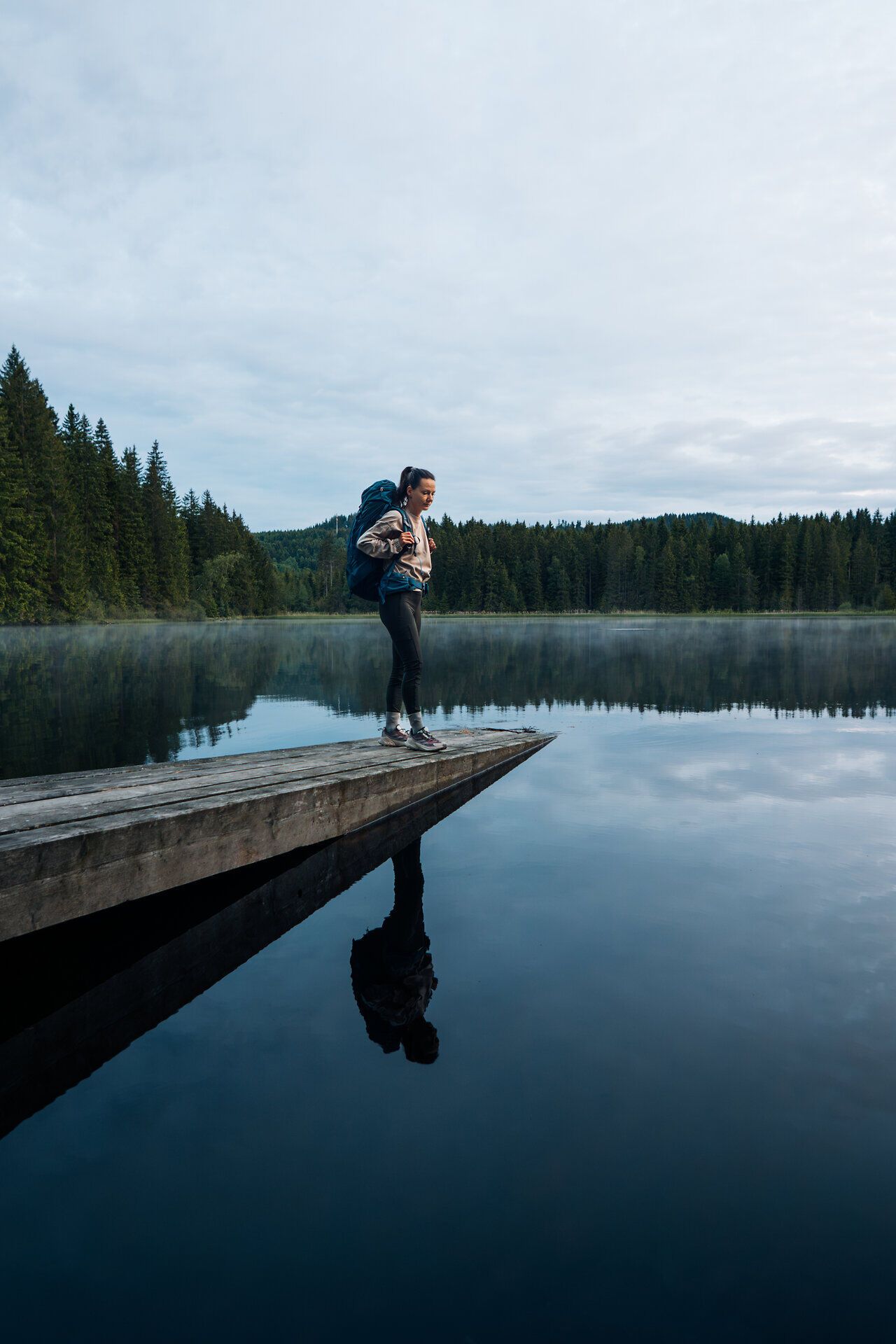 Frau steht auf einem Holzsteg am stillen Stierhübelteich, umgeben von dichter Waldlandschaft im Waldviertel, im Lichterspiel der Dämmerung.
