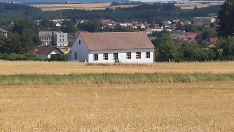 Ein wei&szlig;es Haus mit rotem Dach steht in einem Feld, im Hintergrund eine Stadt und W&auml;lder.