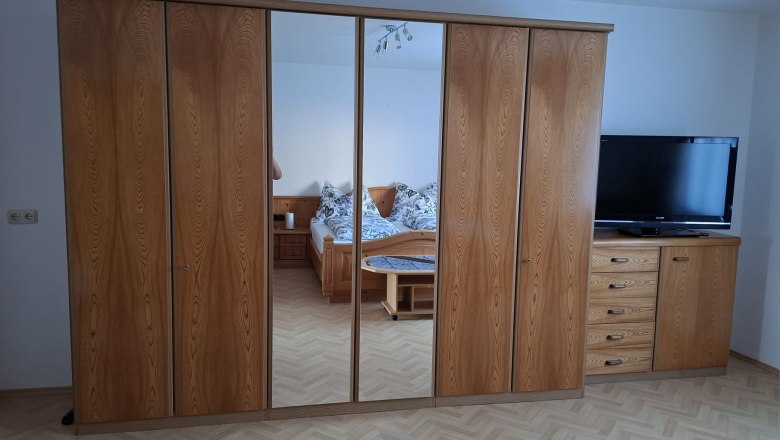 Schlafzimmer mit Holzschrank und Spiegel, Bett im Hintergrund.