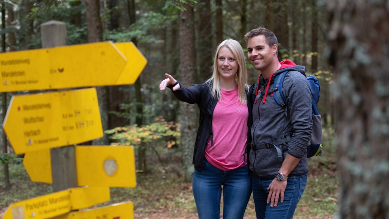 Zwei Personen stehen im Wald neben gelben Wegweisern und schauen in die Ferne.