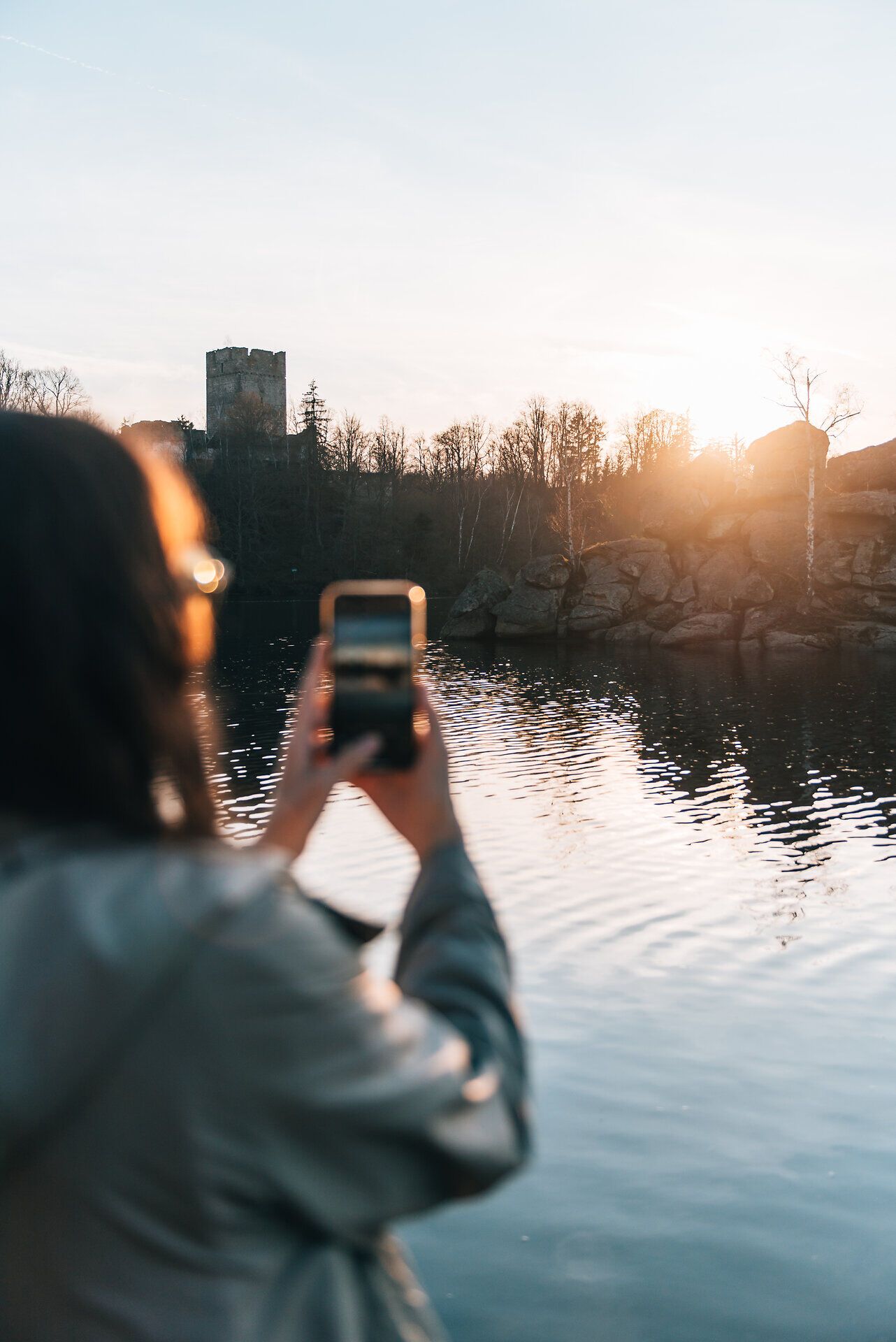 Die sanften Wellen des Wassers spiegeln die warmen Farben des Sonnenuntergangs wider, während eine Person mit ihrem Smartphone den Moment festhält. Umgeben von der ruhigen Natur und dem sanften Licht, entsteht eine harmonische Atmosphäre, die zum Verweilen einlädt.