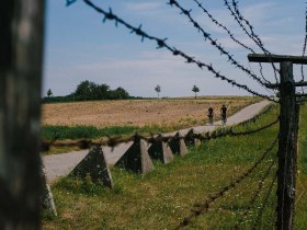Ein malerischer Weg schl&auml;ngelt sich durch die sanften H&uuml;gel, umgeben von &uuml;ppigen Wiesen und dem sanften Rauschen des Windes. Die Stacheldrahtz&auml;une erinnern an vergangene Zeiten und verleihen der Landschaft eine besondere Atmosph&auml;re. Hier, wo Natur und Geschichte aufeinandertreffen, l&auml;dt die Umgebung zum Entdecken und Verweilen ein.